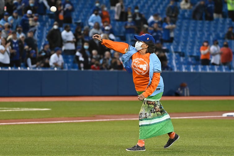 A woman wearing a face mask and in an orange shirt throws a baseball in a large stadium.