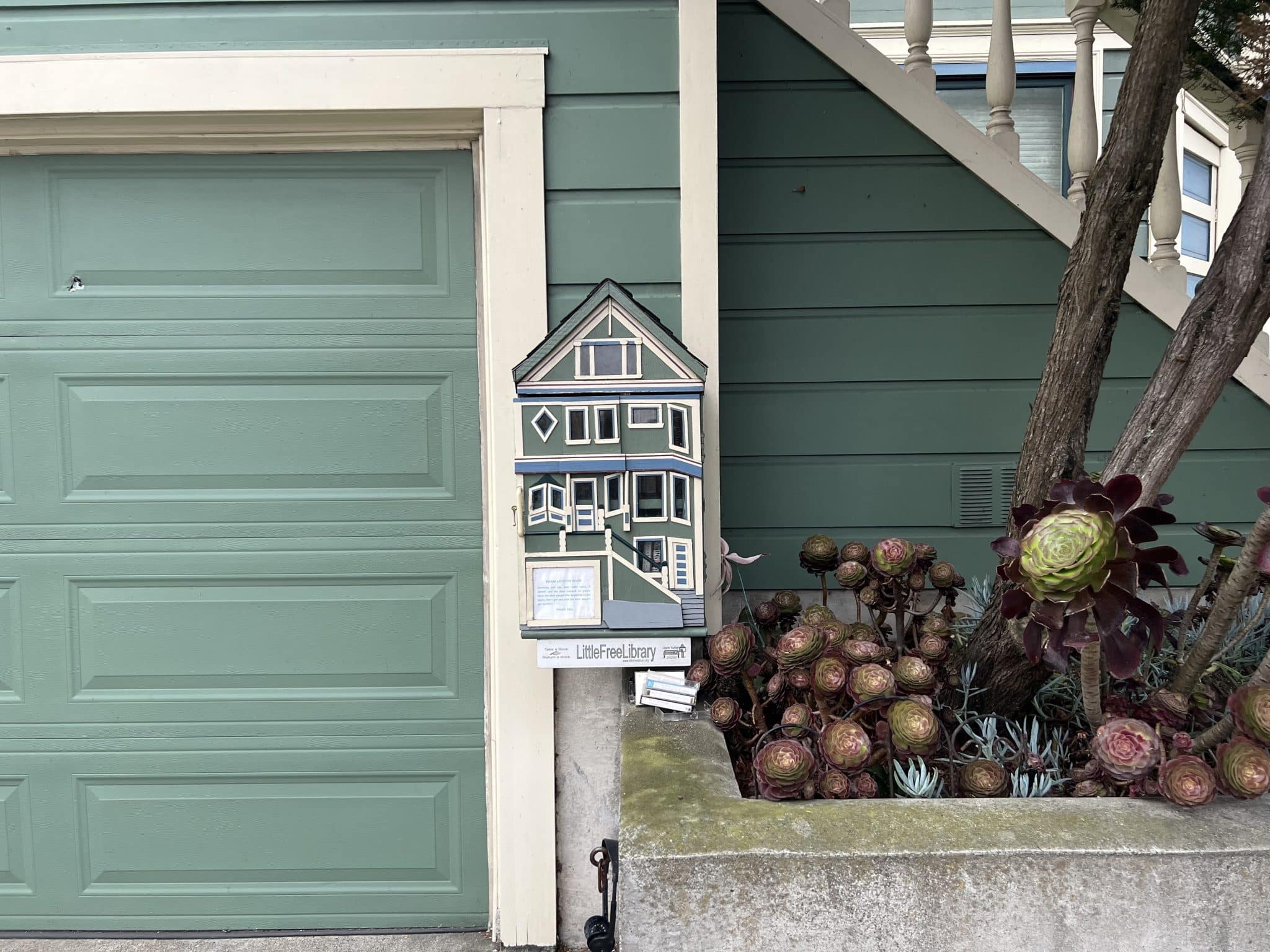 A Little Free Library in the shape of a Victorian house. Painted the same sage green color as the house behind it. 