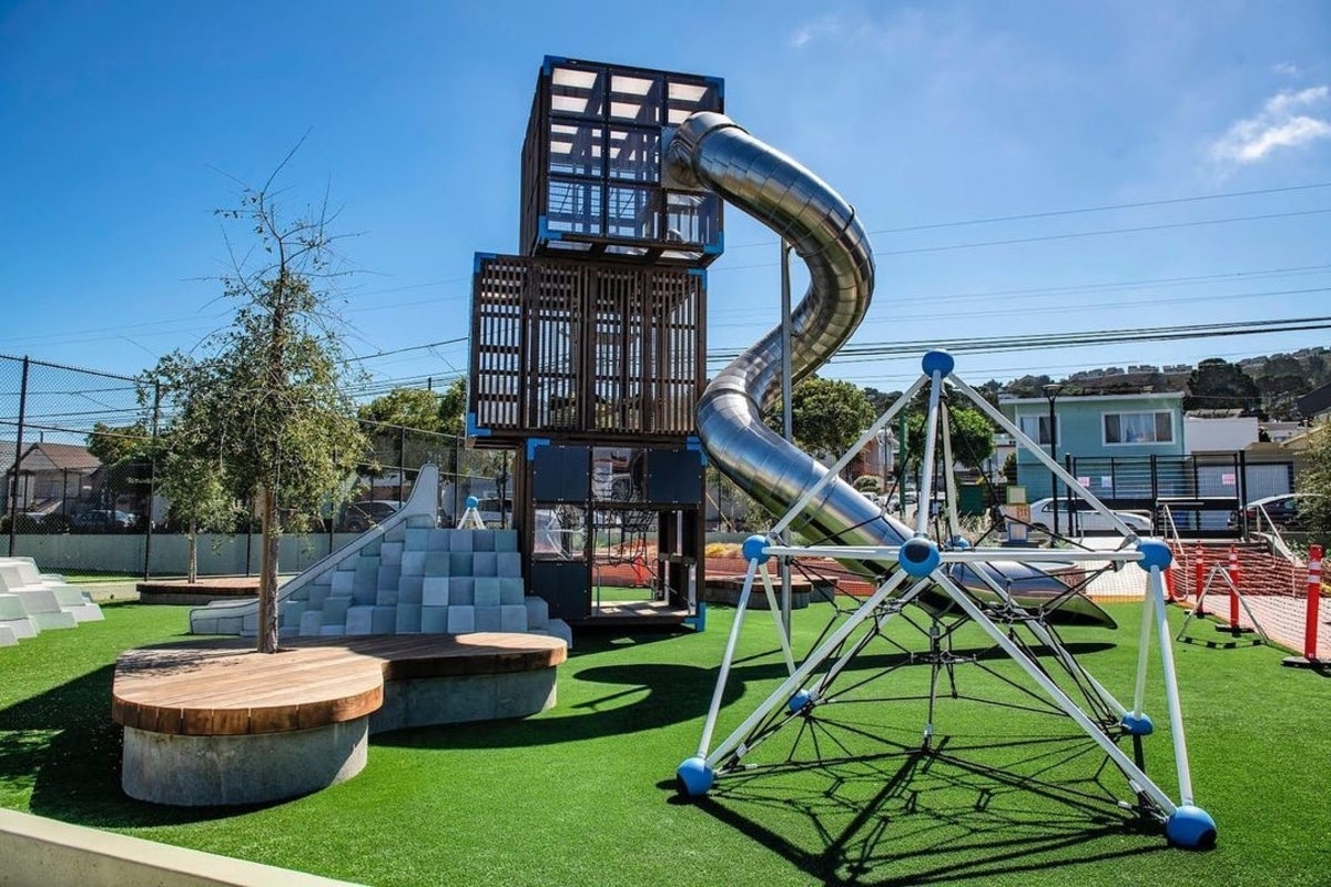 Alice Chalmers Park in San Francisco, featuring a large climbable structure made of stacked transparent boxes with brown frames and a large circular metal tube slide spiraling down.