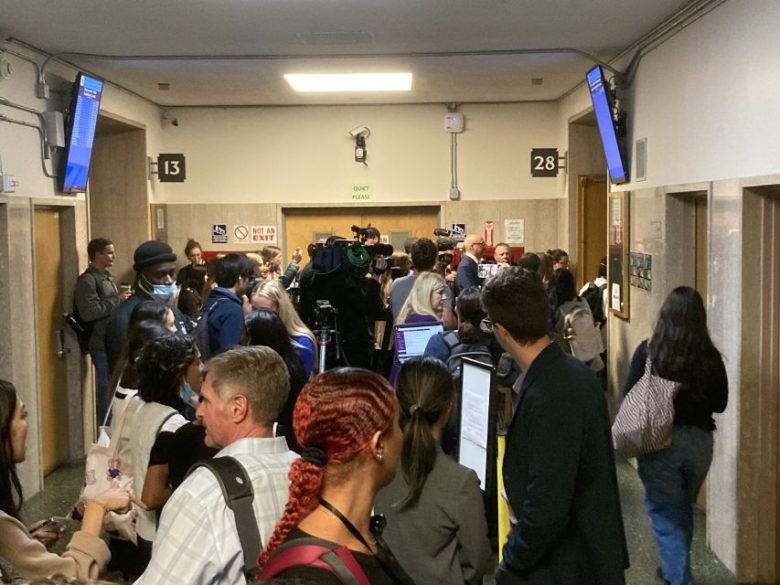 A crowded hallway with people gathered around a camera crew. Signs for room 13 and room 28 are visible on the walls.