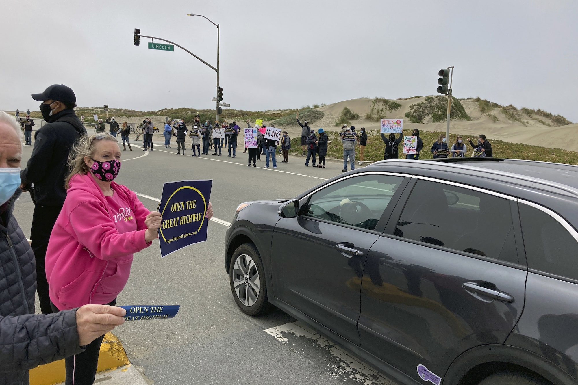 A woman holds a sign that syas 'open the great freeway' to passing motorists near a beachside road