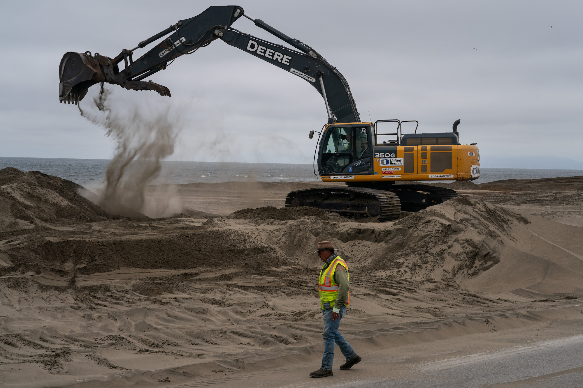 An excavator scoops sand from a stretch of land next to a coastal highway while a person in construction gear walks by