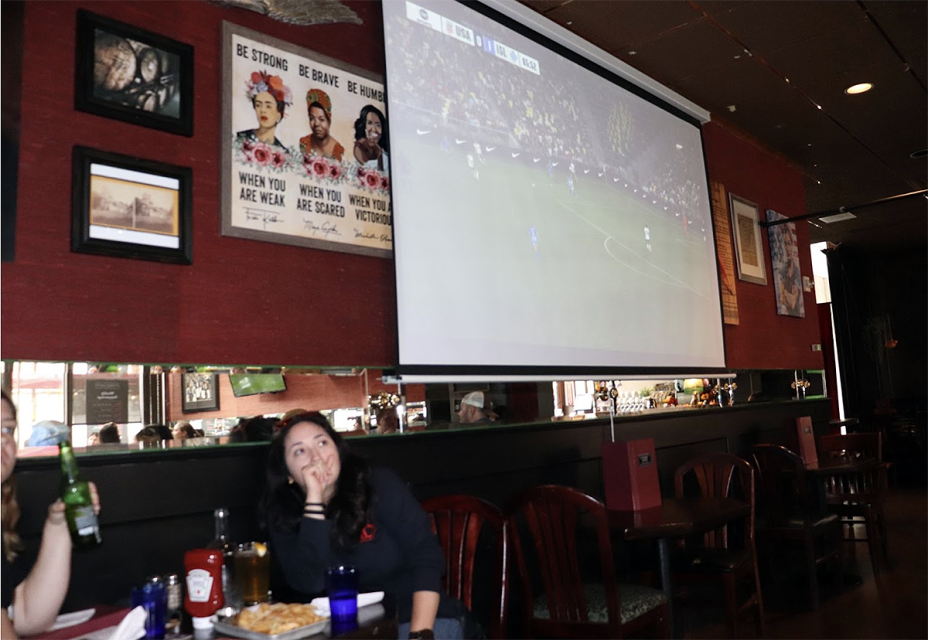 A woman sitting at a table watching a soccer game on a large screen in a restaurant.