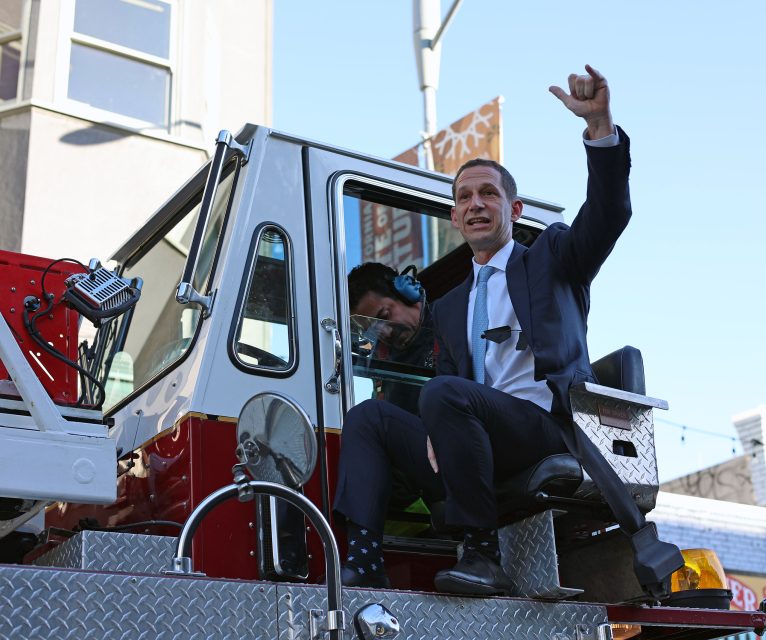 A man in a suit sits on a fire truck, gesturing enthusiastically, while another person is seated inside. The background includes buildings and a streetlight.